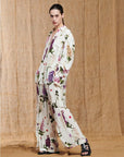 A woman poses against a tan backdrop, wearing HIGH’s "Initiate"—a loose, cream pantsuit with a purple, pink, and green botanical print—paired with black sandals. Her hair is slicked back and her hand is in her pocket.