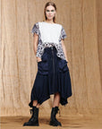 A woman with straight blond hair wears the Uphold navy cargo skirt by HIGH, featuring gathered hems and an elasticated waistband, styled with a white lace-trimmed top and black combat boots, standing before a brown fabric backdrop.