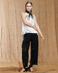 A woman with straight hair and bold green eye makeup models HIGH's Kick-Start tech embroidered jacquard lace pants with cutouts, a white sleeveless blouse with black abstract patterns, and black shoes against a tan fabric backdrop.
