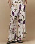 A person wears Dreamer wide-leg silk palazzo pants by HIGH, featuring a bold floral print in purple, red, and green. They have their hands in the pockets and wear black open-toe sandals against a beige background.