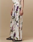 A woman models the Dreamer floral silk palazzo pants by HIGH with black sandals and a sleeveless white top, standing before a beige backdrop.