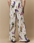 A person stands facing away wearing HIGH's Dreamer luxury women's cream wide-leg silk palazzo pants with a colorful floral print—paired with black sandals—against a plain light brown background.