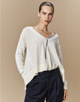 Woman with slicked-back hair wears HIGH's Academy loose cream merino wool V-neck sweater over a black-and-white striped top and high-waisted navy trousers, standing against a beige background.