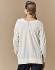 A person with long braided hair is seen from behind wearing HIGH's Academy—a loose, cream crochet knit V-neck sweater with decorative stitching—paired with dark pants against a beige background.