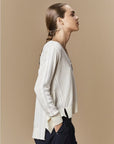 A person with long brown hair in a low braid is shown in profile wearing the cream Academy crochet knit sweater by HIGH and dark pants, standing against a beige background.