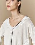 A woman with light skin and light brown braided hair wears the HIGH Academy cream merinos wool V-neck sweater with black trim. Only her head and upper torso are visible against a beige background.