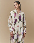Against a beige background, a woman wears HIGH’s "Initiate" set—an oversized tunic and pants with a purple, white, and yellow botanical floral print. Her dark hair is styled in a sleek braid.