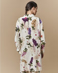 A person with braided dark hair is seen from behind, wearing the HIGH "Initiate" oversized tunic featuring a botanical floral print, posed against a plain beige background.