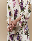 A person models the HIGH "Initiate" cream botanical floral pajama set, designed as an oversized tunic, against a beige background with hands gently clasped. The face is not shown.
