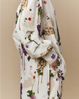 A person stands sideways with hands in pockets, wearing the HIGH "Initiate" oversized tunic in cream, featuring botanical floral prints on a neutral beige background.
