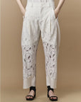 A person wearing HIGH’s Kick-Start tailored, high-waisted pants with abstract patterns and solid panels stands against a beige background, hands in pockets, styled with black sandals and ankle socks.