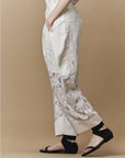 A person stands against a beige background, wearing HIGH's Kick-Start wide leg lace pants with abstract patterns, black sandals, and a white top. Their hands are in their pockets, and only the lower half of their body is visible.