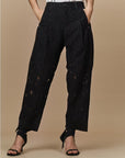 A person wears HIGH's Kick-Start tech embroidered jacquard lace pants with textured, semi-sheer panels, hands in pockets, styled with black strappy heels against a neutral beige background.