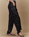 A person models HIGH’s Kick-Start black wide leg lace pants with satin details and sheer patterns, paired with black strappy heeled sandals, posing against a beige background with one hand in their pocket.