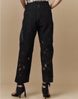 A person stands facing away, wearing HIGH's Kick-Start tailored wide leg lace pants with sheer panels and black cuffs, paired with black ankle-tie high-heeled sandals. The background is plain beige.