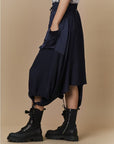 A person stands against a beige background, wearing the HIGH Uphold dark cargo skirt with elasticated waistband, pockets, and drawstring hems, paired with black lace-up combat boots. Only the lower half of the body is visible.