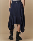 A person stands against a beige background, wearing HIGH’s Uphold navy blue tech satin skirt with an elasticated waistband, gathered drawstring hem, pockets, and black combat boots. The skirt is longer at the sides and back; their hands are in their pockets.