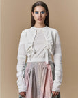A woman with long brown hair models HIGH's "Absolutely"—a white seamless knit cardigan over a white top and pastel plaid pleated skirt—standing against a beige background and looking directly at the camera.