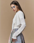 A woman with long brown hair and bold blue eyeshadow models HIGH's Absolutely seamless knit long-sleeve top and pleated skirt with multicolored vertical stripes, standing against a beige background.