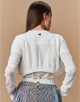 A woman faces away from the camera wearing HIGH's "Absolutely" seamless white knit long-sleeve crop sweater and a high-waisted striped skirt with a white belt, set against a plain beige background.