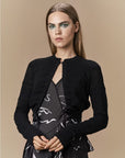 A woman with long straight hair wears the "Absolutely" black fitted cardigan by HIGH over a black dress with abstract white patterns. She stands against a beige background with bold green eyeshadow.