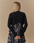 A woman with long blonde hair, seen from behind, wears the HIGH 'Absolutely' fitted black cropped cardigan and a black skirt with white abstract lines against a plain beige background.