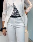 A woman stands indoors in sophisticated style, wearing white pants, a black-and-white patterned blouse, and the HIGH Affair Jacket with an asymmetrical zipper. She completes her look with a black watch and belt.