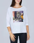 A woman is smiling while wearing SUZI ROHER’s “The Bailey” white long-sleeve shirt with a graphic and the text: "It was great. I love the coffee with charm and such. Coffee In Bed was great! Not love." She stands against a plain background.