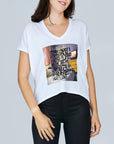 A woman wears SUZI ROHER’s The Velvet tee—a white, boxy-cut shirt with a vibrant Coffee In Bed graphic and black handwritten text: It feels great to love and be loved with charm and cheer. Styled here with black pants.