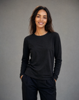 A woman with long dark hair, wearing The CATHERINE Tee By You by SUZI ROHER—a luxurious black crew neck long-sleeve shirt—paired with dark pants, stands smiling with her hands in her pockets against a gray background.