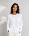A woman with long dark hair, dressed in The CATHERINE Tee By You by SUZI ROHER—a luxury crew neck white long-sleeve shirt—paired with light pants, stands against a plain light background, smiling with her hands in her pockets.