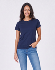 A woman with long, wavy dark hair smiles while standing against a light background. She wears The KARLIE Tee By You in navy blue by SUZI ROHER and blue jeans, with one hand in her pocket.