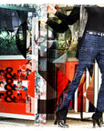 Wearing blue plaid pants, black boots, a black jacket, and SUZI ROHER’s Daily Planet modal silk scarf made in Italy, a person stands by a red phone booth covered in photos and posters. A large ampersand graphic overlays the image.