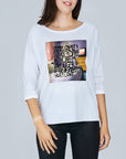 A woman smiles in front of a gray background, wearing The Bailey, a luxurious white long-sleeve shirt by SUZI ROHER featuring a graphic print and black text: "It’s great! I love the coffee with charm and sugar! It’s great! Not love!.
