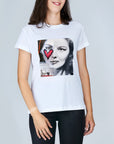 A woman with shoulder-length brown hair smiles in SUZI ROHER's The Niki tee, a classic 100% cotton white shirt featuring a black-and-white Audrey Hepburn portrait partly covered by a red heart and abstract designs.