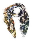 The Cottage by SUZI ROHER is a silk scarf with frayed edges, arranged in a loose spiral on a white background. Made in Italy, it features an abstract pattern in green, yellow, blue, and brown.