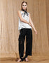 A woman with straight hair and bold green eye makeup models HIGH's Kick-Start tech embroidered jacquard lace pants with cutouts, a white sleeveless blouse with black abstract patterns, and black shoes against a tan fabric backdrop.