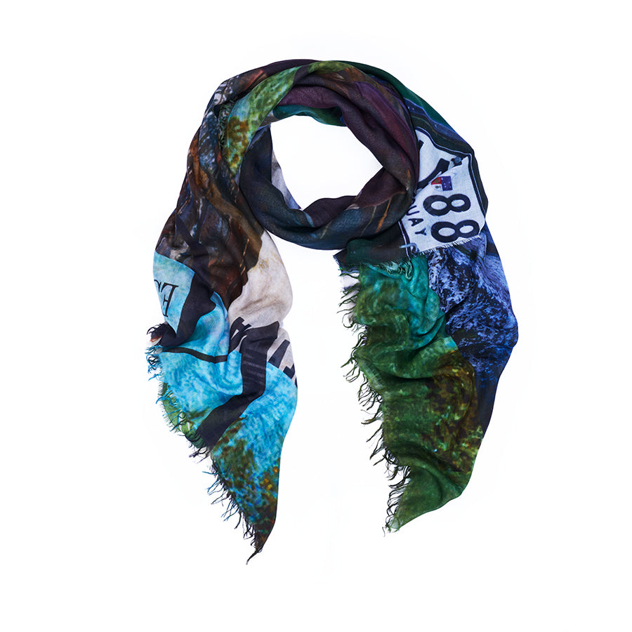 The SUZI ROHER “Spanish Vogue” scarf displays original artwork with a scenic print in blue, green, and white hues, featuring a visible road sign marked 88. Digitally printed for vibrant detail and finished with frayed edges, this colorful accessory is shown on a white background.