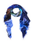 A square scarf from SUZI ROHER, named “Sunset Blvd.,” features frayed edges and showcases a vibrant blue and multicolored abstract pattern inspired by original artwork. Digitally printed, the scarf is draped in a loose loop against a white background.