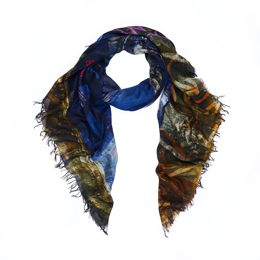 The “Against All Odds” scarf by SUZI ROHER features a vibrant digital print in dark blue, brown, yellow, and touches of purple, with frayed edges and is styled in a relaxed loop against a white background.