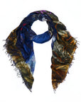 The “Against All Odds” scarf by SUZI ROHER features a vibrant digital print in dark blue, brown, yellow, and touches of purple, with frayed edges and is styled in a relaxed loop against a white background.