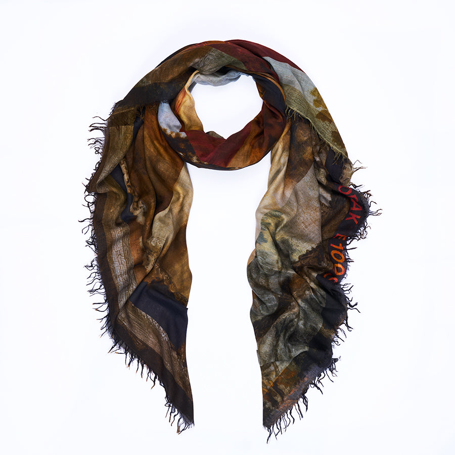 Presenting the “Da Vinci” scarf by SUZI ROHER—a lightweight, rectangular accessory with frayed edges. Digitally printed for sharp detail, this scarf showcases an abstract design in earthy browns, blacks, and subtle muted hues. Red text appears partially on the right side, adding unique flair to its artful look. Here, it is styled in a loose loop for a relaxed finish.