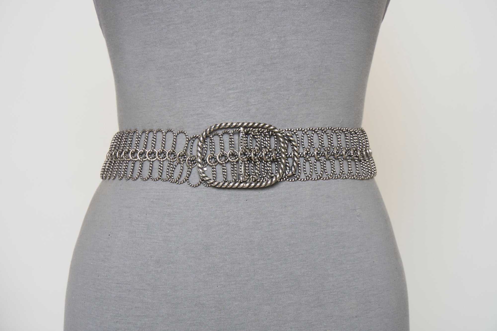 A mannequin showcases SUZI ROHER's 6826—a silver metallic chain belt with a large oval buckle—styled over a fitted gray dress.
