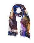 The SUZI ROHER "Brave Hearted" scarf, featuring bold blue, purple, yellow, brown, and green abstract patterns with frayed ends, is shown in a loose loop on a white background—perfect as a cozy accessory for fall.