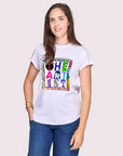 A smiling woman with long brown hair wears SUZI ROHER's The Karlie—a white T-shirt with a colorful THE ARTIST graphic—paired with blue jeans, standing against a light, plain background.