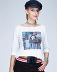 A woman poses confidently against a light background wearing The Bailey white boxy cut tee by SUZI ROHER, paired with a navy cap, colorful belt, and silver jewelry. One hand rests on her hip as she looks directly at the camera.