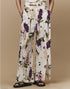 A person wears Dreamer wide-leg silk palazzo pants by HIGH, featuring a bold floral print in purple, red, and green. They have their hands in the pockets and wear black open-toe sandals against a beige background.