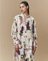 Against a beige background, a woman wears HIGH’s "Initiate" set—an oversized tunic and pants with a purple, white, and yellow botanical floral print. Her dark hair is styled in a sleek braid.