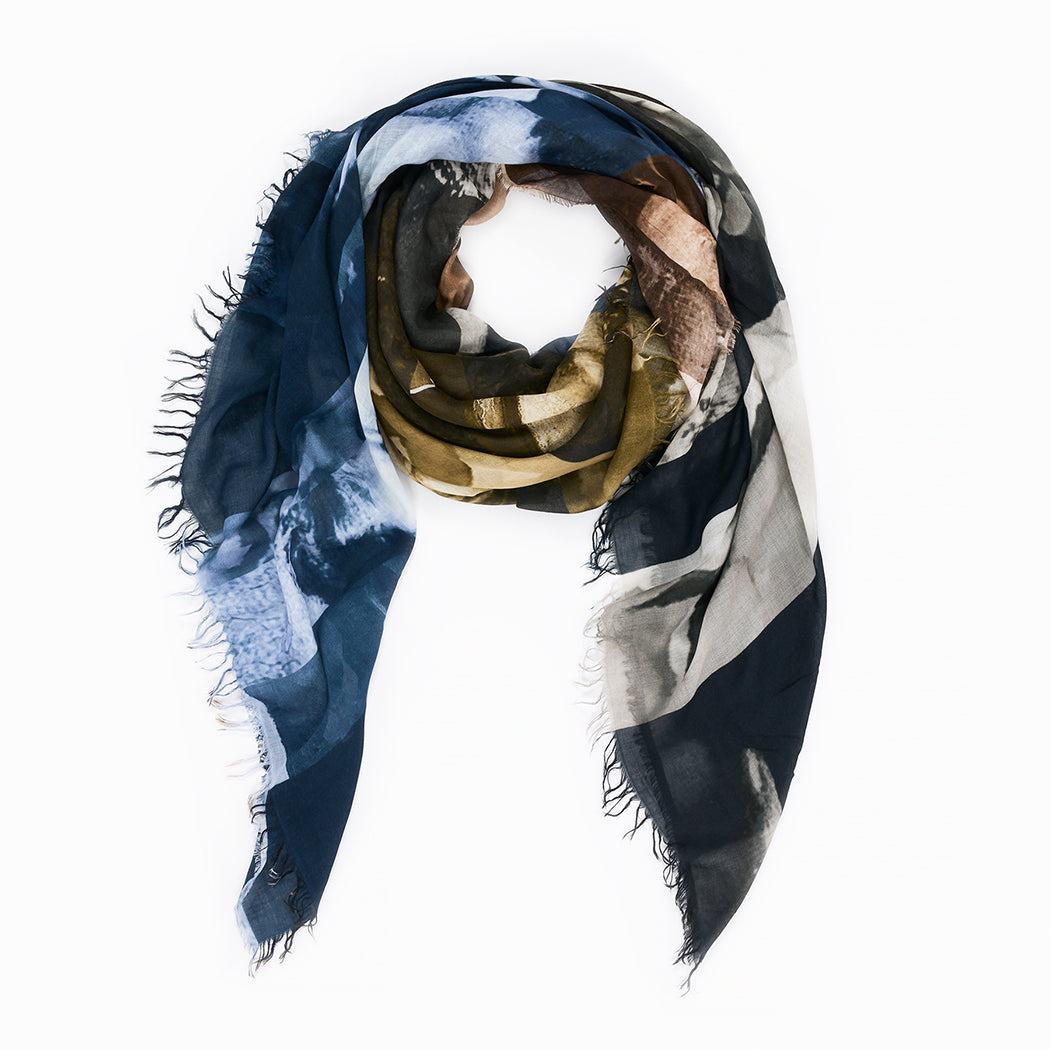 Euphoria by SUZI ROHER is a square-shaped, multi-colored scarf with fringed edges, showcasing digitally printed original artwork in abstract patterns of dark blue, olive green, brown, and cream. The scarf is loosely arranged in a loop on a white background.