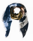 Euphoria by SUZI ROHER is a square-shaped, multi-colored scarf with fringed edges, showcasing digitally printed original artwork in abstract patterns of dark blue, olive green, brown, and cream. The scarf is loosely arranged in a loop on a white background.